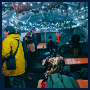A photograph of an indoor event at Plattfields Market Garden. The event is taking place in their sustainably sourced polytunnel with fairy lights and people sat at picnic benches